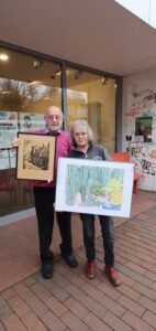 Zwei Besucher zeigen ihre ersteigerten Bilder, ein Holzschnitt, der eine Häuserreihe in Kappeln darstellt und ein Aquarell, das Kakteen zeigt.