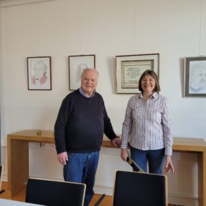 Ilse Demasius vor 4 ihrer Zeichnungen bei der Hängung der Bilder. Sie trägt eine gestreifte Bluse, daneben ein älterer Mann im blauen Pullover.