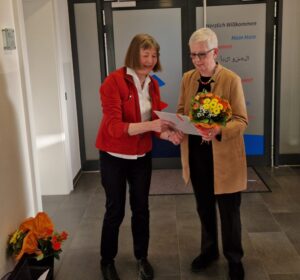 Ilse Demasius in roter Jacke übernimmt einen Blumenstrauß von Inke Asmussen in senfgelben Longjacket.