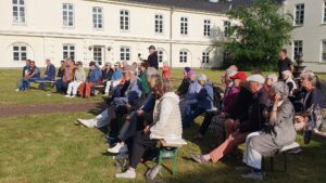 Zahlreiche Besucher sitzen auf Biergartenbänken im Halbrund im Innenhof. Die Sonne scheint.