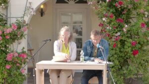 Eine Frau und ein Mann an einm Tisch sitzend halten eine Lesung ab.