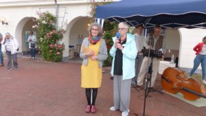 Zwei Frauen vor einem blauen Zelt unter dem zwei Männer stehen und ein Kontrabass liegt.