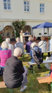 Gäste der Veranstaltung sitzen vor dem Schauspielerpaar und hören sich die Lesung an. Die Sonne scheint.