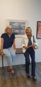 Zwei Frauen auf einem Tisch sitzend vor einem Meeresbild von Elke Schady im Gespräch.