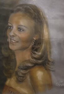 Ein Portrait einer jungen hübschen blonden Frau in Pastellkreide gearbeitet in braun und goldtönen vor grauem Hintergrund von Elke Schady.