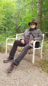 Arten Eden 2025 (11) Foto: M. Roos Ein Mann mit Steppjacke und Hut sitzt auf einer weißen Gartenbank. Hinter ihm Wald,
