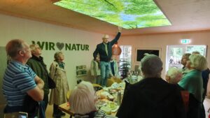 Arten Eden 2025 (12) Foto: M. Roos Thorsten Roos mit einer Gruppe von Besuchern erklärt die Deckenbeleuchtung im GreenHaus. Er steht auf einm Stuhl und zeigt nach oben.
