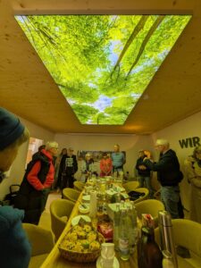 Arten Eden 2025 (30) Foto: H.P. Junge Eine Gruppe von Besuchern steht rund um einen gedeckten Tisch. An der Decke ein rechteckiges Foto von Bäumen, das mit Licht hinterlegt wurde.