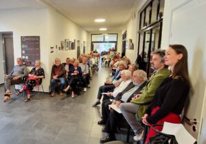 Ein Blick ins Foyer des Quartiers 36, wo die Gäste den Ansprachen lausche. Der Flur ist voll gefüllt mit Gästen, auf Stühlen sitzend.