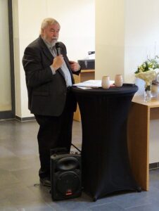 Herr Meyer hält die Laudatio. Er trägt einen dunkelgrauen Anzug und hält ein Mikrofon.