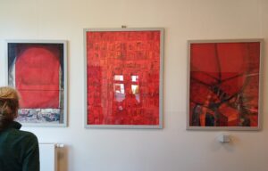 3 abstrakte Gemälde, Hochformat, gerahmt in Rottönen. Das linke ist etwas realistischer und bildet einen Blick aus dem Fenster, das durch eine rotes Rollo dreiviertel verdeckt ist.