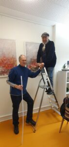 Bernd Wilms auf der Leiter und Volker Neumann mit Zollstock daneben beim Bilder hängen.