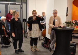 Janine Wilhöft hält ein Mikrofon, daneben ihre Vertretung und Inke Asmussen. Im Hintergrund Musiker im Foyer des Quartier 36.