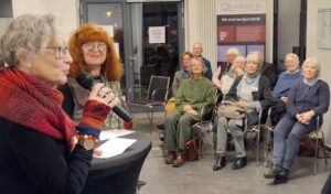 Jutta Heilmann hält eine Ansprach, daneben Heidemarie Utecht, beide vor Gästen der Vernissage.