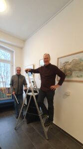 Zwei Männer lächeln in die Kamera im Foyer des Quartier 36. Der vordere steht auf der Leiter.