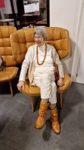 Eine Frau in beige gekleidet in einem gorßen safrangelben Sessel lächelt.