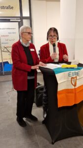 Zwei Frauen in roten Jacken am Stehpult. Eine mit Mikrofon.