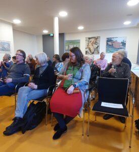 Sie sehen Gäste der Finissage sitzen auf Stuhlreihen, im Hintergrund Gemälde von Claus Vahle.