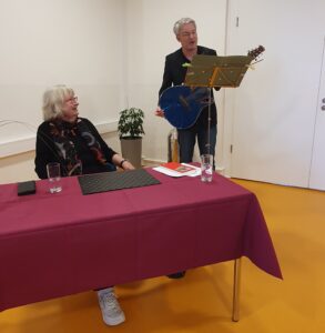 Annegret Vahle am Tisch sitzend schaut zu Patrick Vahle an der Gitarre.
