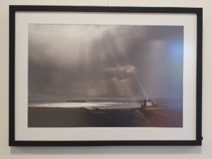 Foto einer kleinen Fischerhütte mit Blick auf das gleißende Meer. Von oben beleuchtet ein Sonnenstrahl das Haus durch eine dicke Wolkendecke hindurch.