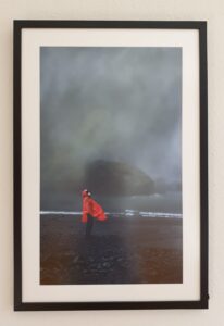 Foto mit einem Mann am Strand vor stürmischer See. Er trägt ein leuchtend oranges Regencape und steht vor einem dunklen Hintergrund.